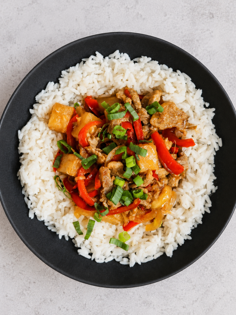 Pineapple pork stir fry with red capsicum and spring onion served over a ring of white rice in a black bowl