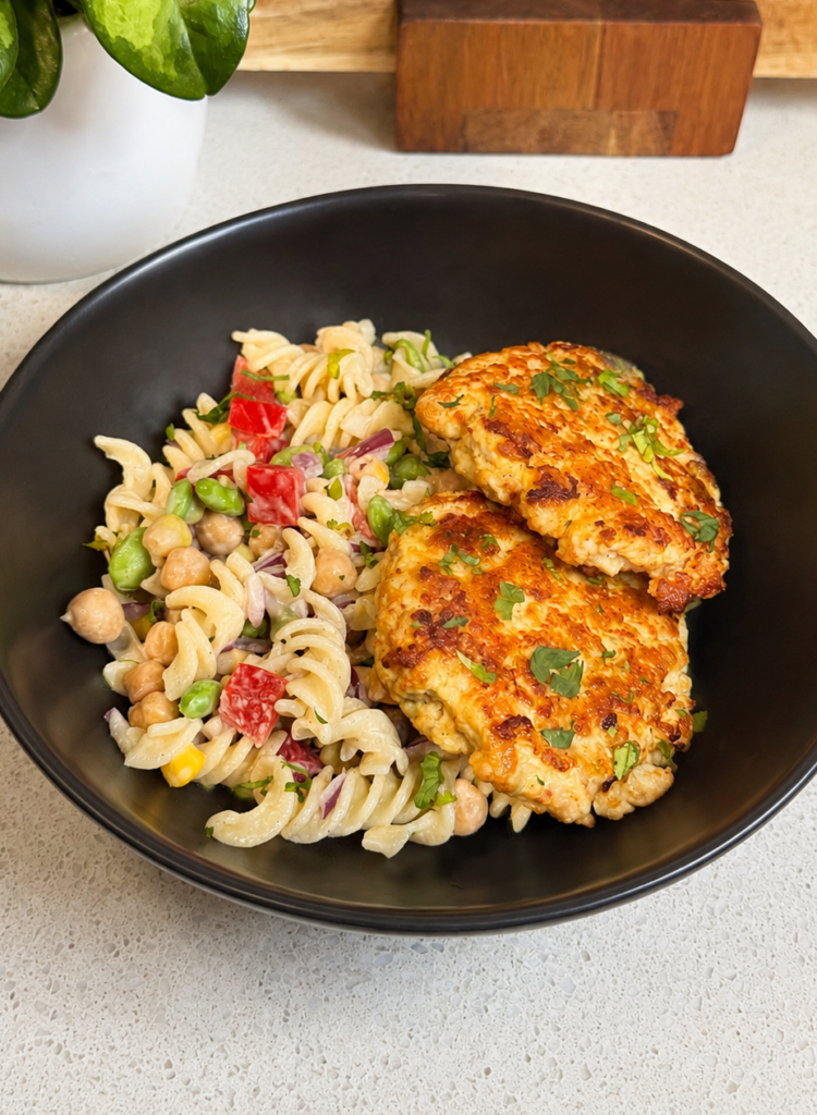 Two smoky chicken rissoles served with a creamy pasta salad of chickpeas, edamame, corn and capsicum in a black bowl